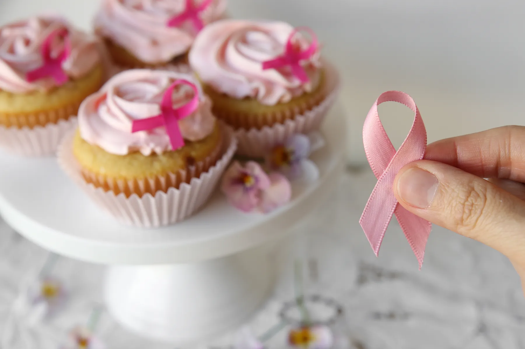 Hand holding a pink awareness ribbon beside pink-iced charity cupcakes